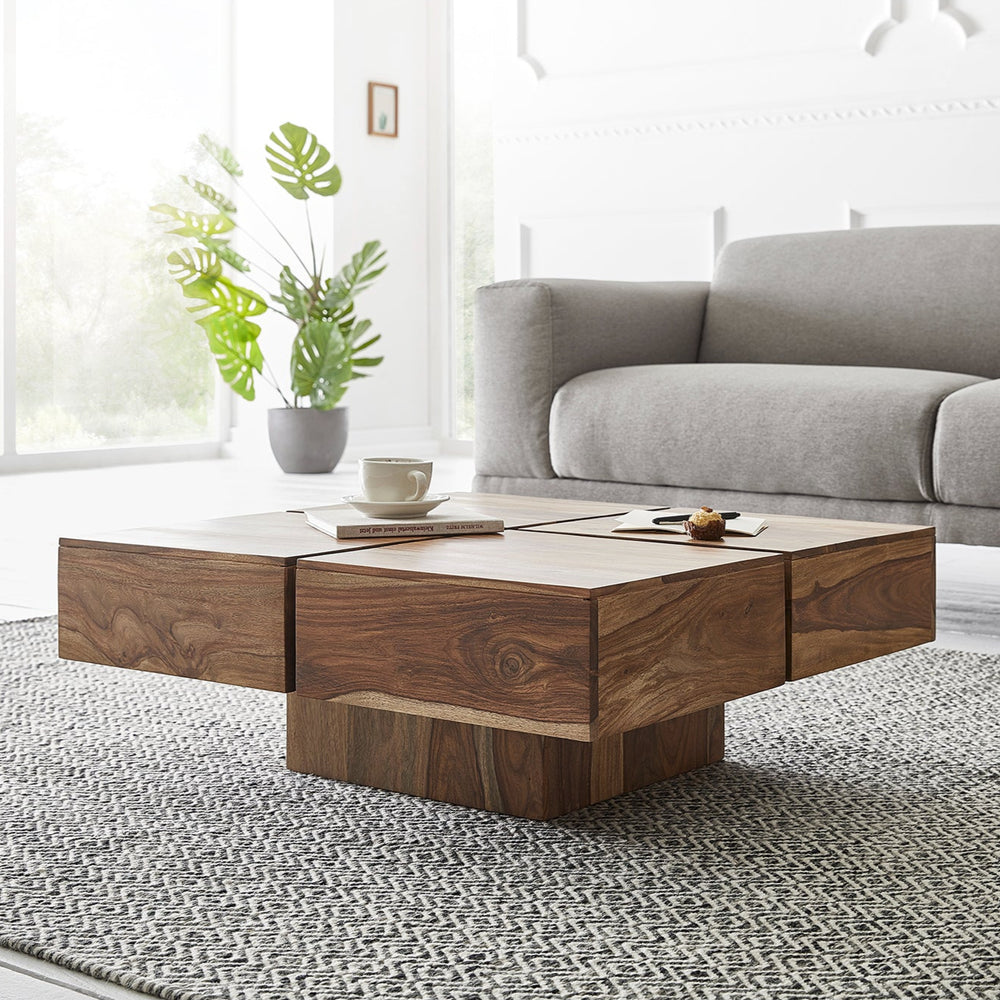 A rectangular wooden coffee table with a natural finish, placed on a grey area rug in a living room setting, with a sofa in the background and a potted plant to the left.
