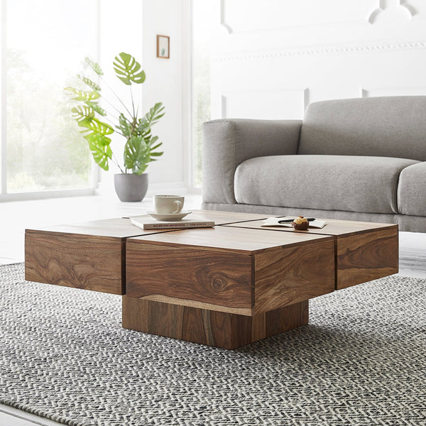 A rectangular wooden coffee table with a natural finish, placed on a grey area rug in a living room setting, with a sofa in the background and a potted plant to the left.